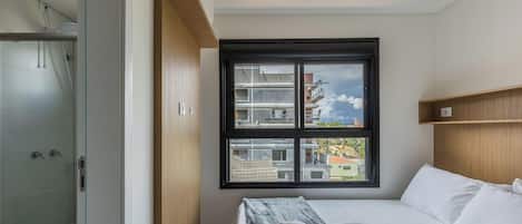 A hotel room with a bed, a window with a view of buildings, a wooden headboard, and a shower area.