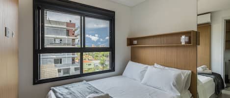 Un dormitorio con un ventanal grande, una cama con ropa de cama blanca, un cabecero de madera y una pequeña estantería de madera.