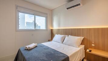 A modern hotel room with a large bed, a bedside table, a window, and an air conditioning unit.