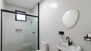 Un baño moderno con ducha envuelta en cristal, un lavabo blanco con grifo negro y un espejo ovalado grande en la pared.