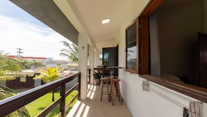A balcony with a wooden railing, a small table, two stools, a potted plant, and a view of a green lawn and buildings. - Apt in Condo w/ Pool 500m From the Beach Enl0002 (Porto Seguro)