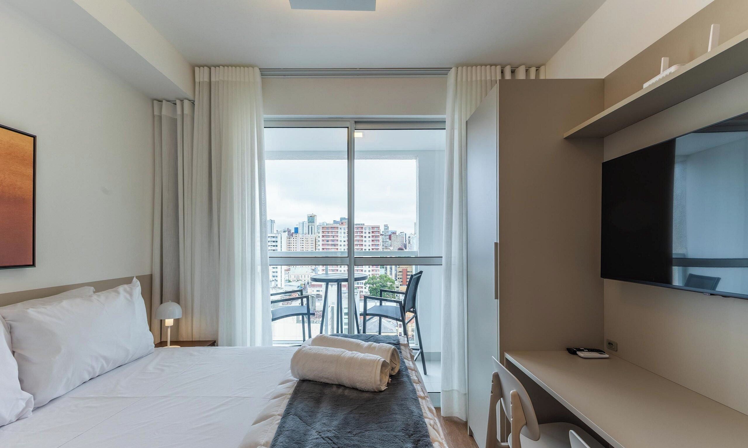 A hotel room with a bed, a desk, a chair, a TV, and a balcony with a city view.