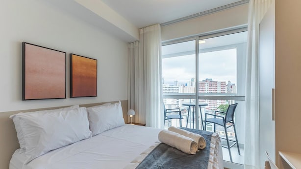 Quarto de hotel com uma cama grande, duas obras de arte emolduradas, uma varanda com vista para a cidade e uma mesa com cadeiras.