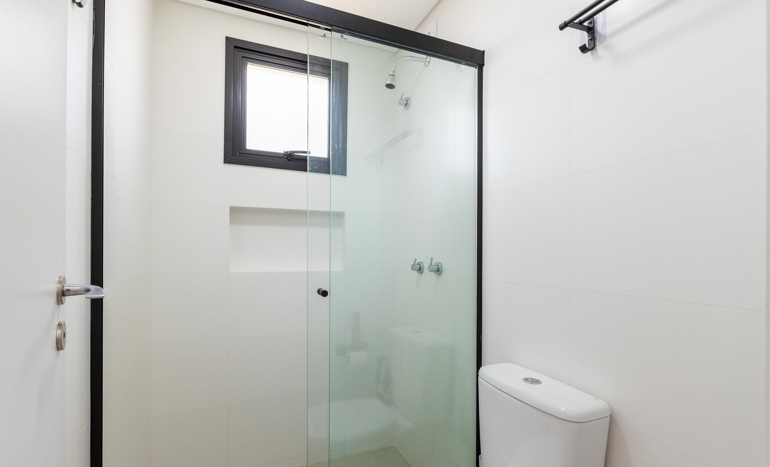 Ducha con cerramiento de vidrio, pequeña ventana, inodoro blanco y puerta de vidrio.