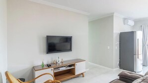 A modern living room with a flat-screen TV mounted on the wall, a wooden entertainment unit, a sofa, and a refrigerator. - Apt in Cond. w Pool, 650m fr/ Beto Carrero Lav0308 (Penha)