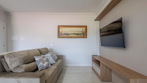 A living room with a beige sofa, patterned cushions, a framed picture on the wall, and a flat-screen TV mounted on the wall. - Apt 650m fr/ Beto Carrero in Cond w/ Pool Lav0401 (Penha)