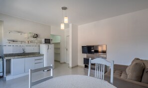 A modern kitchen with white cabinets, a microwave, and a refrigerator. There is a dining area with a white table and chairs, and a sofa in the living room. - Comfortable Apart 200m From the sea Psd202 (Florianópolis)