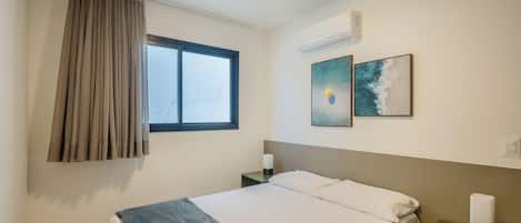 A hotel room with a bed, a window with curtains, an air conditioning unit, and two wall-mounted artworks.