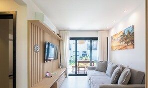 A modern living room with a flat-screen TV mounted on a wooden panel wall, a grey sofa with multiple cushions, and a large window offering a view of the outdoors. - 2 Bedroom Apart in Condominium With Pool Vlp0111 (Mata de São João)