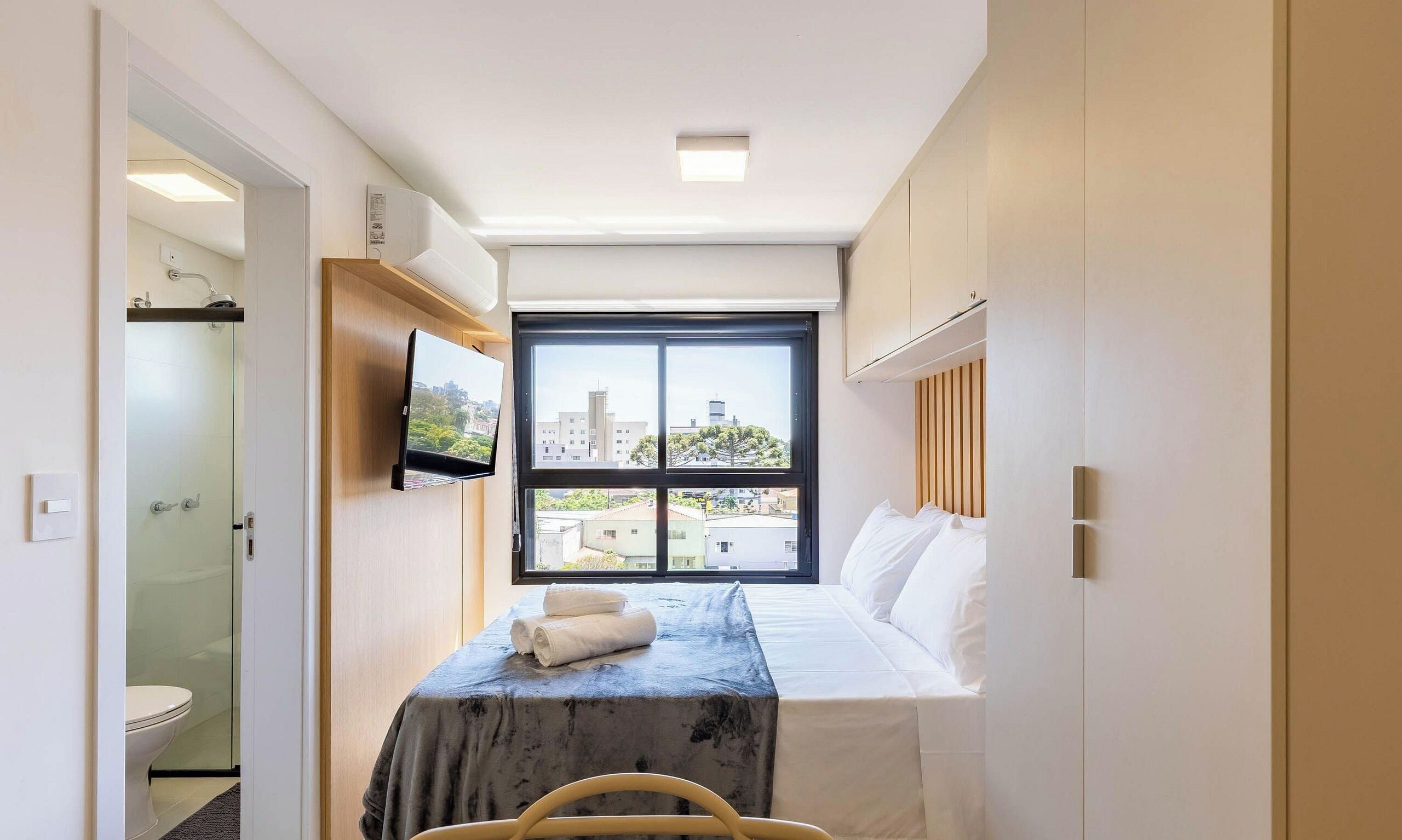 Ein Hotelzimmer mit einem Bett, einem an der Wand montierten Fernseher, einem Fenster mit Blick auf die Stadt und einem Badezimmer mit Duschkabine.