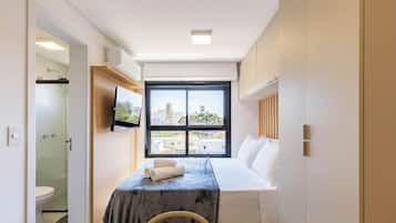 A hotel room with a bed, a TV mounted on the wall, a window with a city view, and a bathroom with a glass shower enclosure.