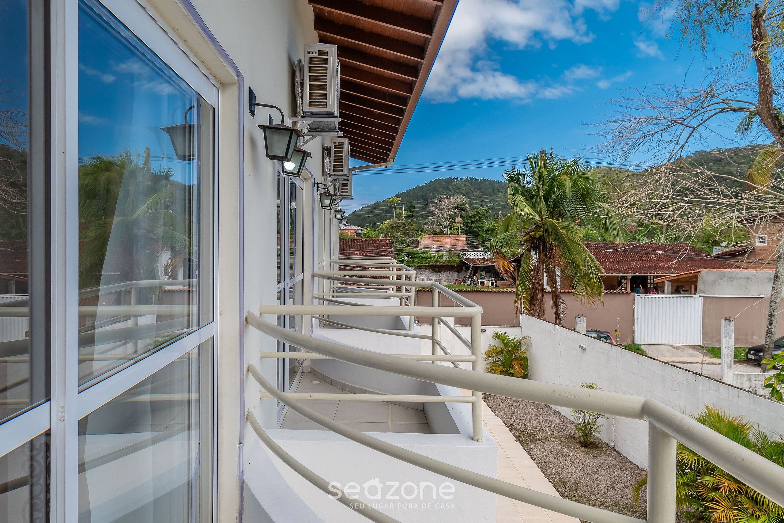 Un edificio moderno con balcón, unidades de aire acondicionado y vistas a una zona residencial con casas y palmeras.