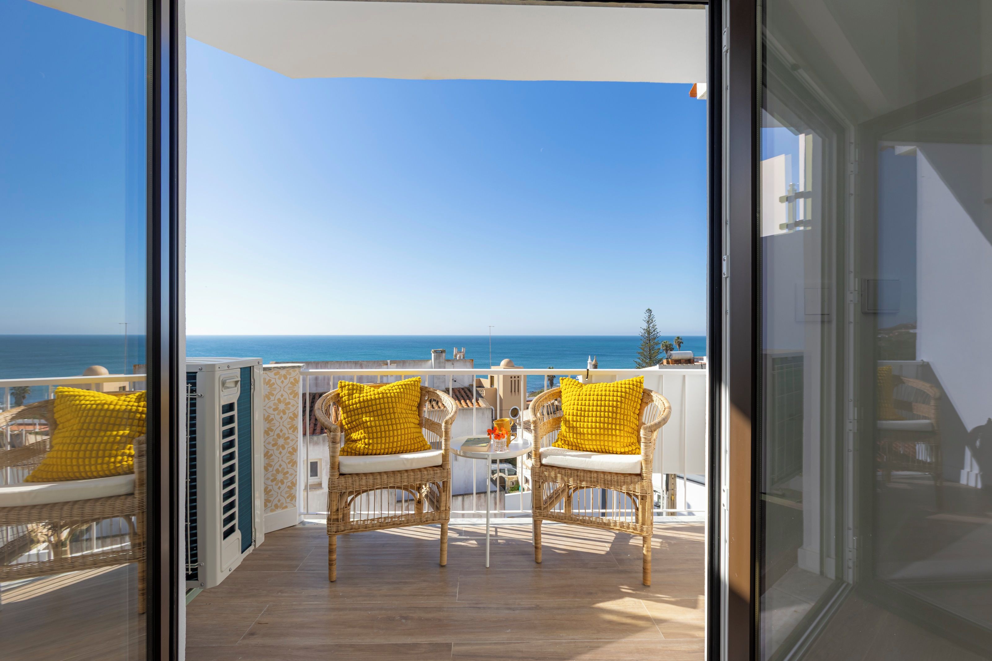A balcony with wicker chairs and yellow cushions, overlooking a clear blue sky and a distant coastline.