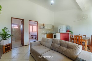 Una sala de estar con un sofá, una mesa de comedor con sillas, una zona de cocina con electrodomésticos y una planta en maceta.