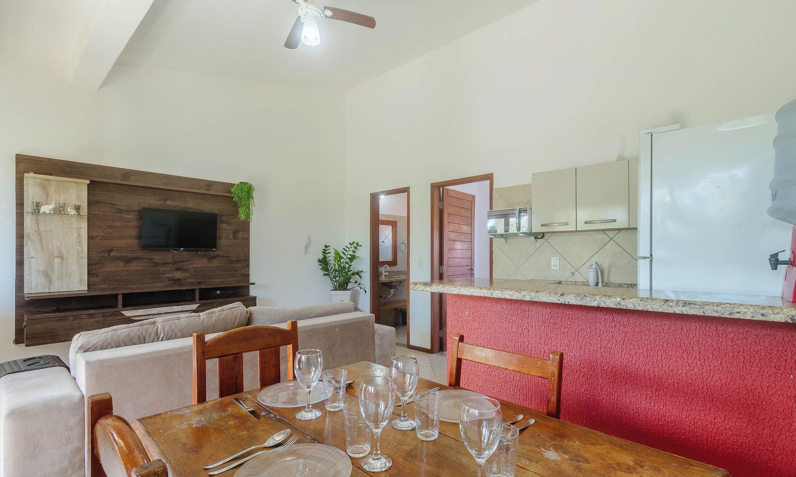 Uma sala de jantar com uma mesa de madeira posta para refeição, uma televisão presa a um painel de madeira, e uma cozinha com geladeira e armários.