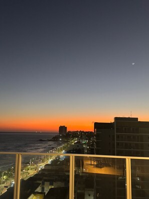 Paisagem urbana costeira ao entardecer, com edifícios e uma estrada que segue para o mar.