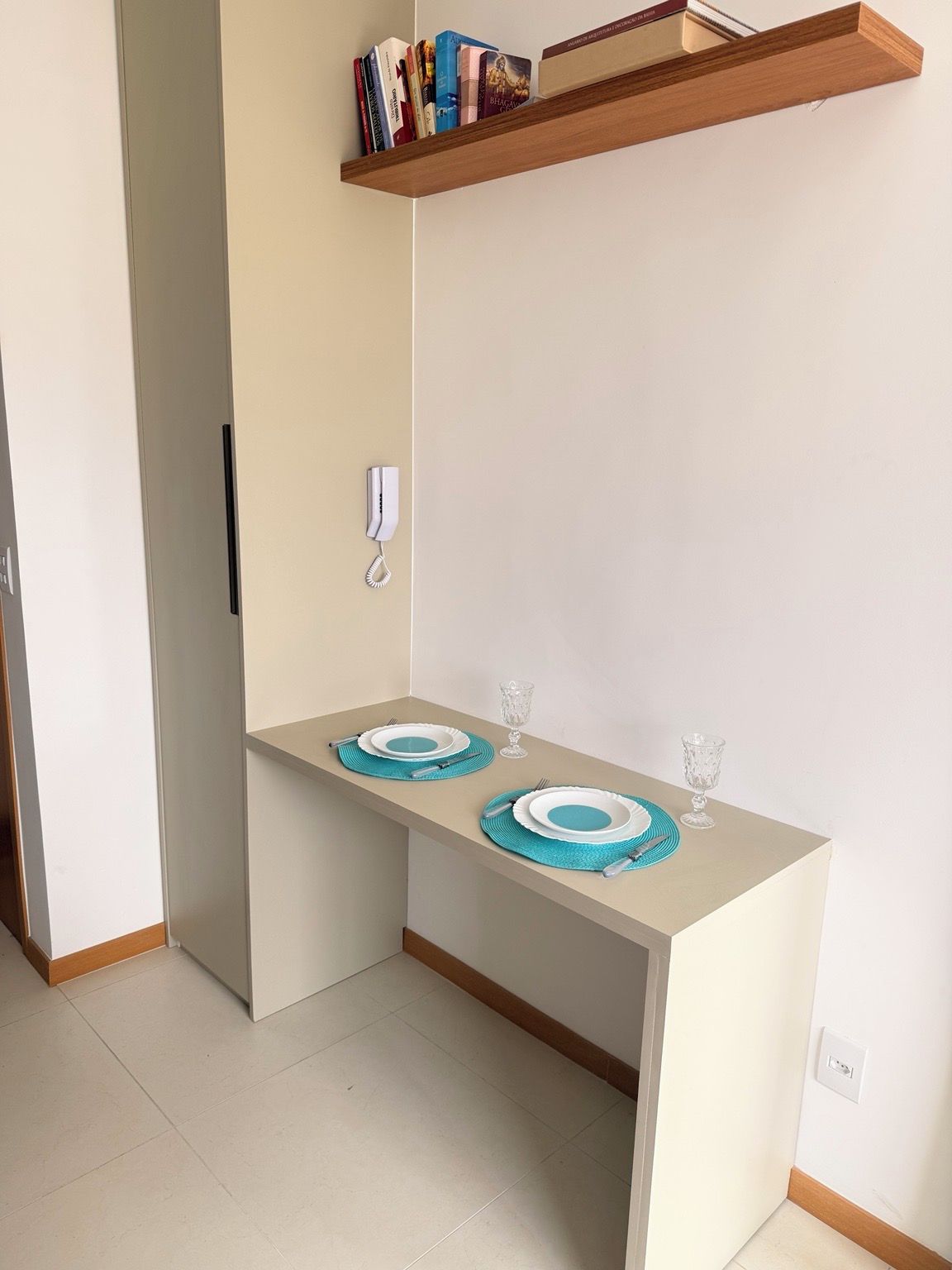 Ein Eckzimmer mit einem kleinen Tisch für zwei Personen, einem Bücherregal darüber und einem Telefon an der Wand.