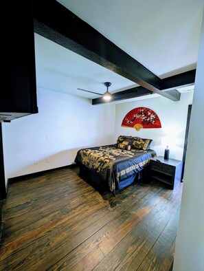Un dormitorio con cama, mesita de noche, ventilador de techo y un abanico decorativo rojo en la pared.