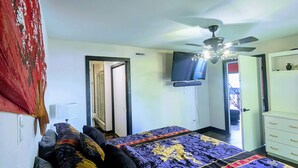 Quarto com cama, ventilador de teto, televisão e porta para outro espaço.