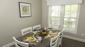 Une salle à manger avec une table mise pour quatre personnes, des chaises, une fenêtre avec des stores et un tableau encadré au mur.