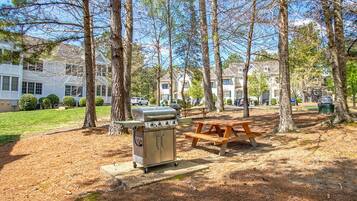 Un espace pique-nique équipé d’un barbecue et de bancs, entouré d’arbres et de bâtiments résidentiels.
