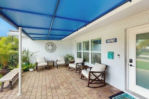 Ein überdachter Patio-Bereich mit Terrazzoböden, weißen Wänden und einem blauen Baldachin. Mehrere Stühle und ein Tisch sind vorhanden, sowie ein dekoratives Wandelement.