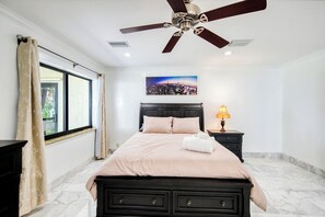 Ein Schlafzimmer mit einem Bett, einem Deckenventilator, einem Nachttisch mit Lampe, einem Fenster mit Vorhängen und einem Stadtbild an der Wand.