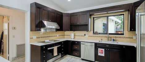 A modern kitchen with dark cabinetry, a stainless steel range hood, a dishwasher, and a window with a view.