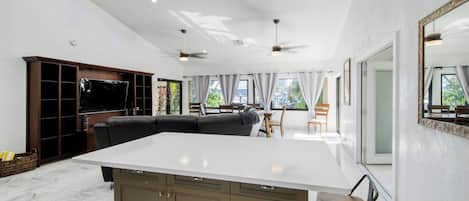 Amplia sala de estar con una isla blanca grande, gabinetes marrón oscuro, un televisor de pantalla plana y ventiladores de techo.