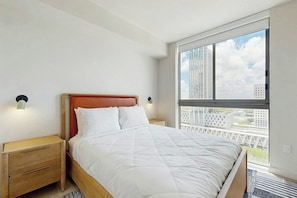 A bedroom with a large window, a bed with white bedding, a wooden headboard, and two bedside tables with lamps. - Luxe in the Sky - Skyline Views (Miami)