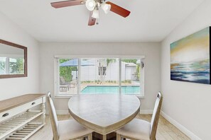 A room with a round table, chairs, a ceiling fan, a window with blinds, and a painting on the wall. - The Front House & Cottage (Fort Lauderdale)