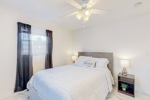 Ein Schlafzimmer mit einem Bett, einem Nachttisch, einem Deckenventilator und einem Fenster mit Vorhängen.