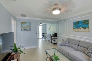 Ein Wohnzimmer mit einer Couch, einem Glastisch und einem Deckenventilator. An der Wand hängt ein Gemälde und auf dem Boden steht eine Pflanze.