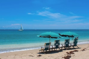 Ein Strand mit zwei grünen Sonnenschirmen und Liegestühlen, auf denen 'O.B.S.' steht.