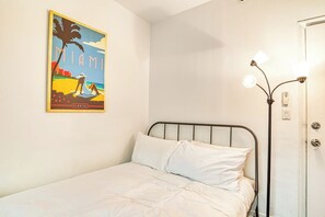 A single bed with white bedding in a room with a white wall and a framed poster of a beach scene. - So South Beach - Waterfront Condo (Miami Beach)