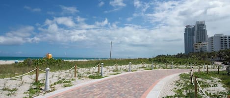 A paved walkway leading to a beach with a lifeguard tower, surrounded by sand dunes and buildings in the background.