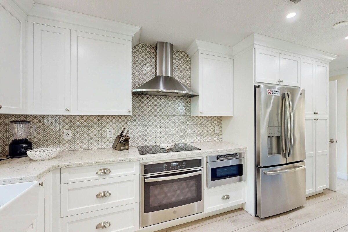 A modern kitchen with white cabinetry, a stainless steel range hood, a built-in oven, a microwave, and a refrigerator.