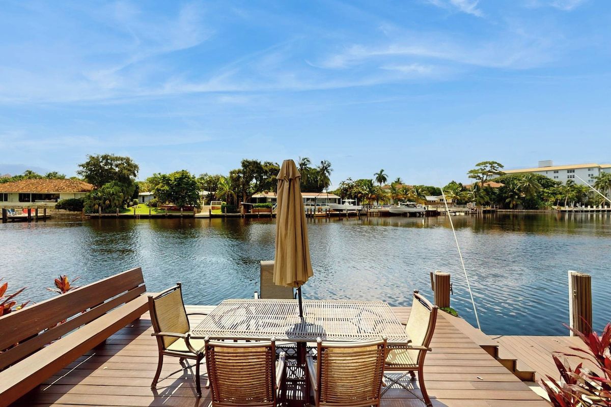 A wooden deck with a table and chairs overlooking a calm body of water, with buildings and trees in the background.