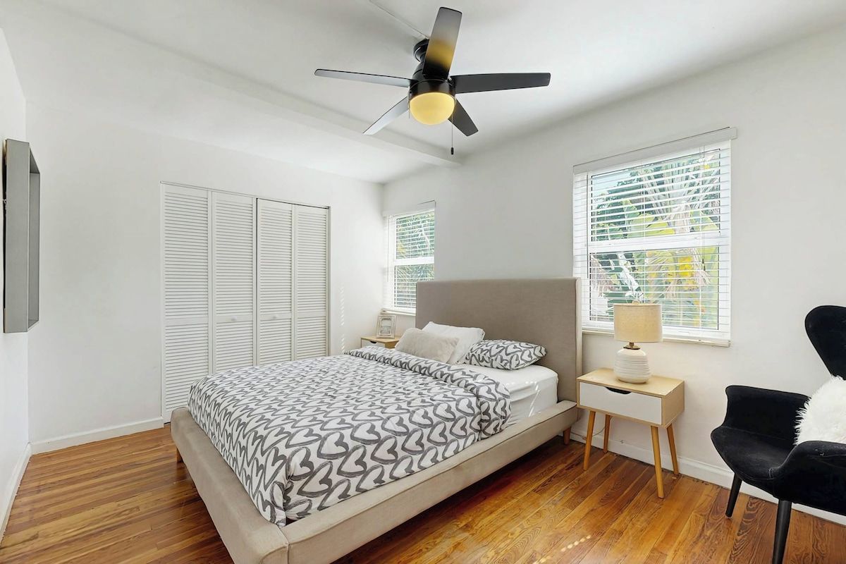 Un dormitorio con cama, mesitas de noche, una silla, ventilador de techo y ventana con persianas.