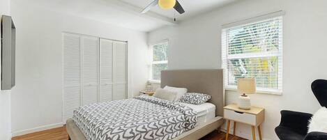 Un dormitorio con cama, mesitas de noche, una silla, ventilador de techo y ventana con persianas.