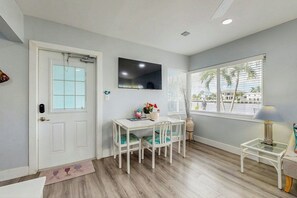A room with a white door, a flat-screen TV mounted on the wall, a dining table with chairs, a small side table, and a window with blinds. - Aloha Beach Holiday - Waterfront (Pompano Beach)