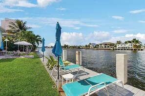A waterfront hotel with a poolside area featuring lounge chairs and umbrellas, a grassy lawn, and a view of the water and distant buildings. - Aloha Beach Holiday - Waterfront (Pompano Beach)