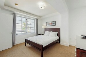 A bedroom with a bed, a window with curtains, a bedside table, and a wall-mounted light fixture. - The Carlyle 3G - Near Beach (Miami Beach)