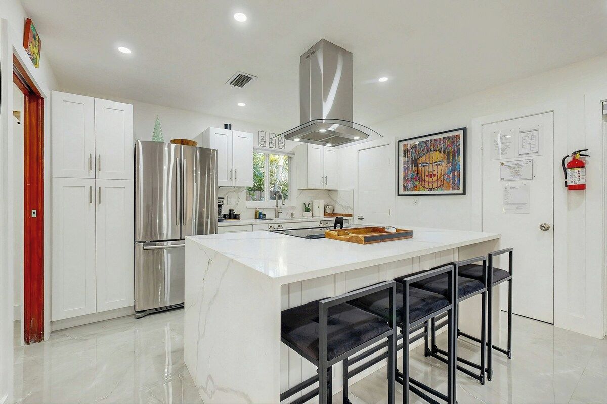 Een moderne keuken met witte kasten, een roestvrijstalen koelkast, een centraal eiland met barstoelen, en een brandblusser aan de muur.