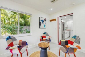 A modern living room with two colorful chairs, a round wooden table, and a large window with a view of greenery. - 2 BR Home with Private Patio & Grill (Wilton Manors)