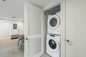 A laundry room with two stacked front-loading washing machines, a door with a louvered panel, and a small seating area with a table and chairs. - The Grand Vista 4501 (Miami)