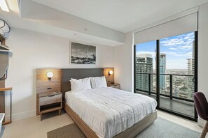 Quarto moderno com cama grande, duas mesas de cabeceira, um criado-mudo, janela com vista para a cidade e um quadro na parede.