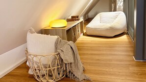 Un dormitori tipus loft acollidor amb sostre inclinat, una taula de nit amb una làmpada, una cadira de vimet i un seient de plàstic.