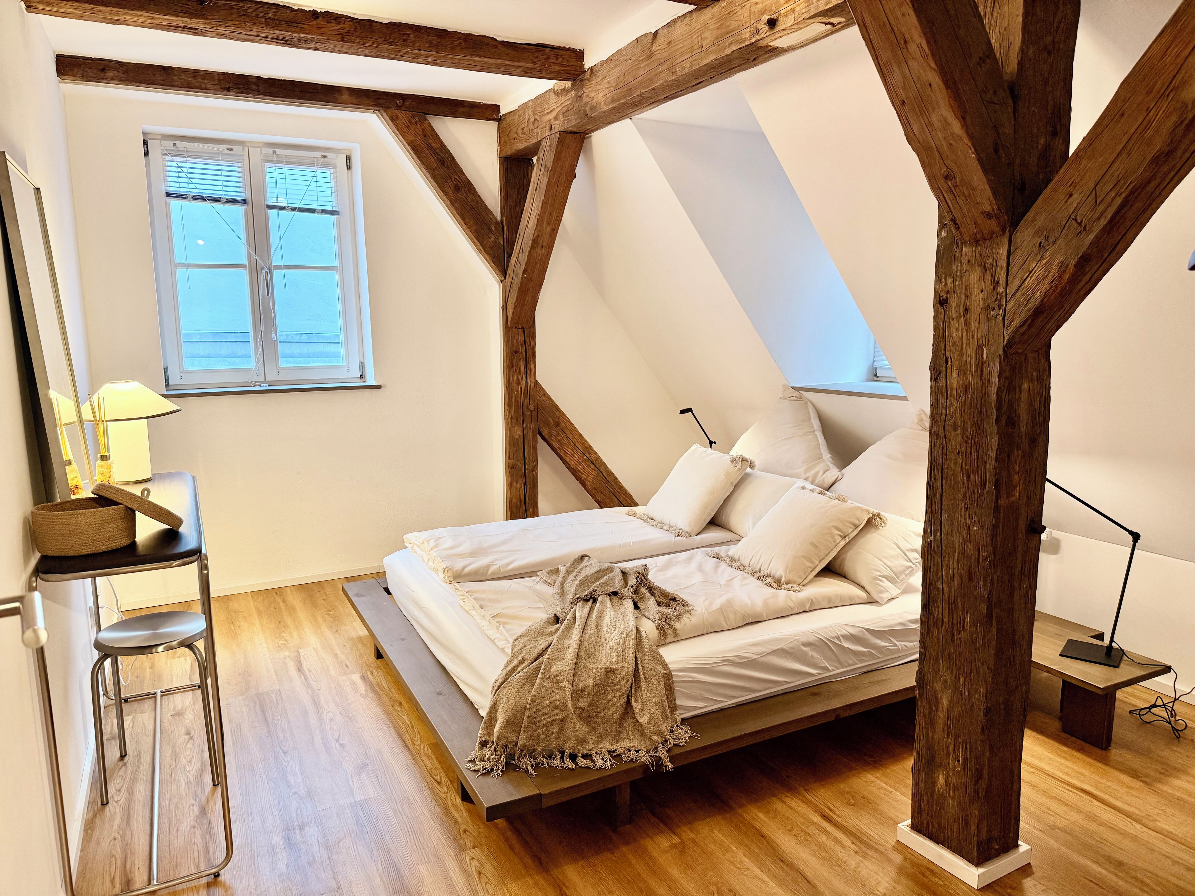 Ein Schlafzimmer mit geneigter Decke, Holzbalken, einem Bett mit weißer Bettwäsche, einem Schreibtisch mit Lampe und einem Fenster mit Jalousien.