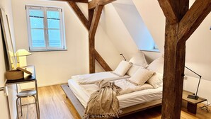 A bedroom with a sloped ceiling, wooden beams, a bed with white bedding, a desk with a lamp, and a window with blinds. - A Hidden Gem in the Heart of Ingolstadt (Ingolstadt)